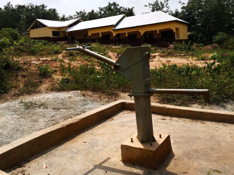 A hand water pump in the foreground with a school building behind